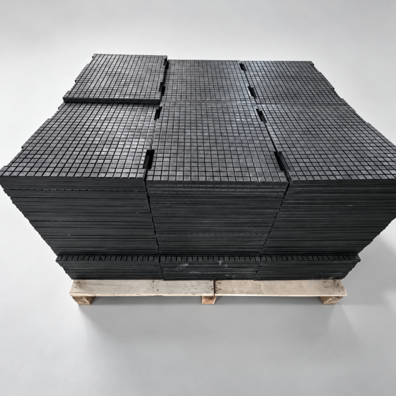 iksonic-composite-ceramic-liner-panels-pallet Stacked dark IKSONIC composite ceramic liner panels with square-grid surfaces and edge slots on a wooden pallet.