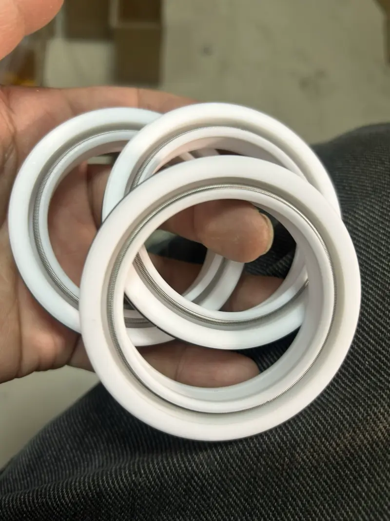 A hand holding three white annular shaft seals with visible circumferential springs.