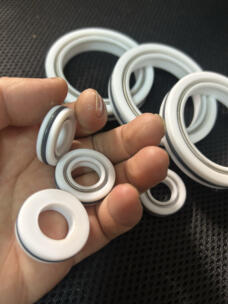 Hand holding three small white shaft seals with different faces and the outer dark sealing ring visible.
